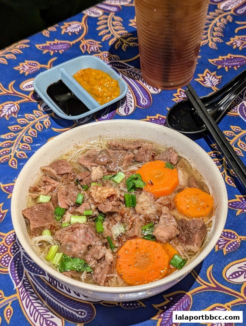 A hearty bowl of beef noodle soup with carrots and green onions, served with dipping sauces.