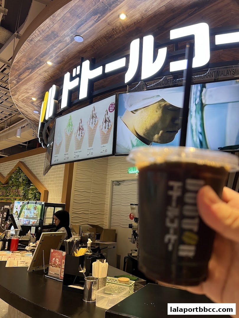 A person holds a coffee cup in front of a DOUTOR COFFEE shop counter with ice cream displays.