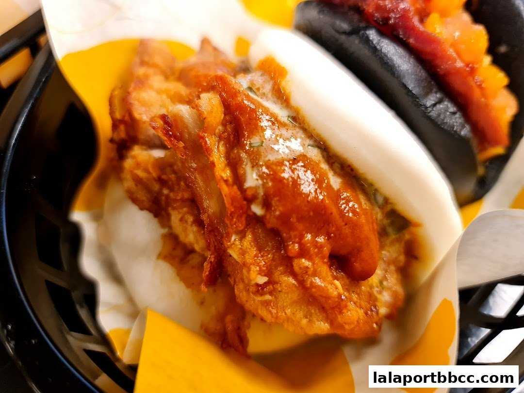 Close-up of two delicious bao buns, one with crispy fried chicken and sauce, the other with a dark bun and topping.