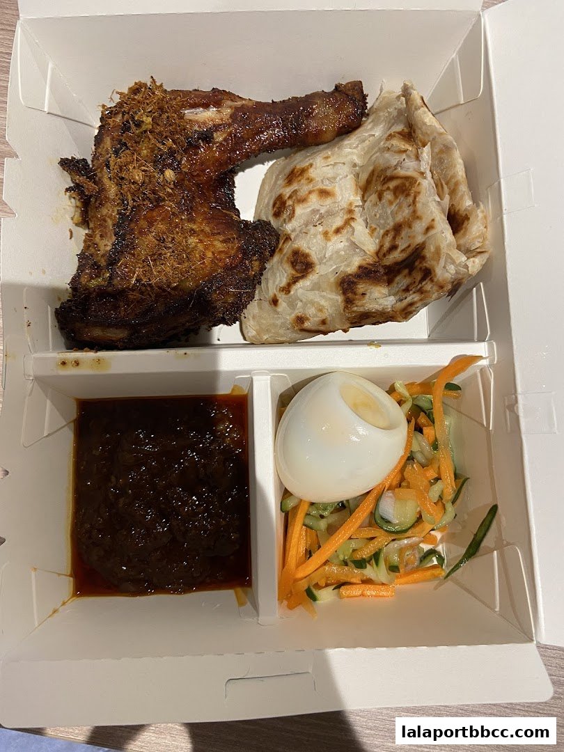 A takeaway box containing fried chicken, roti, sambal, and a side salad with boiled egg.