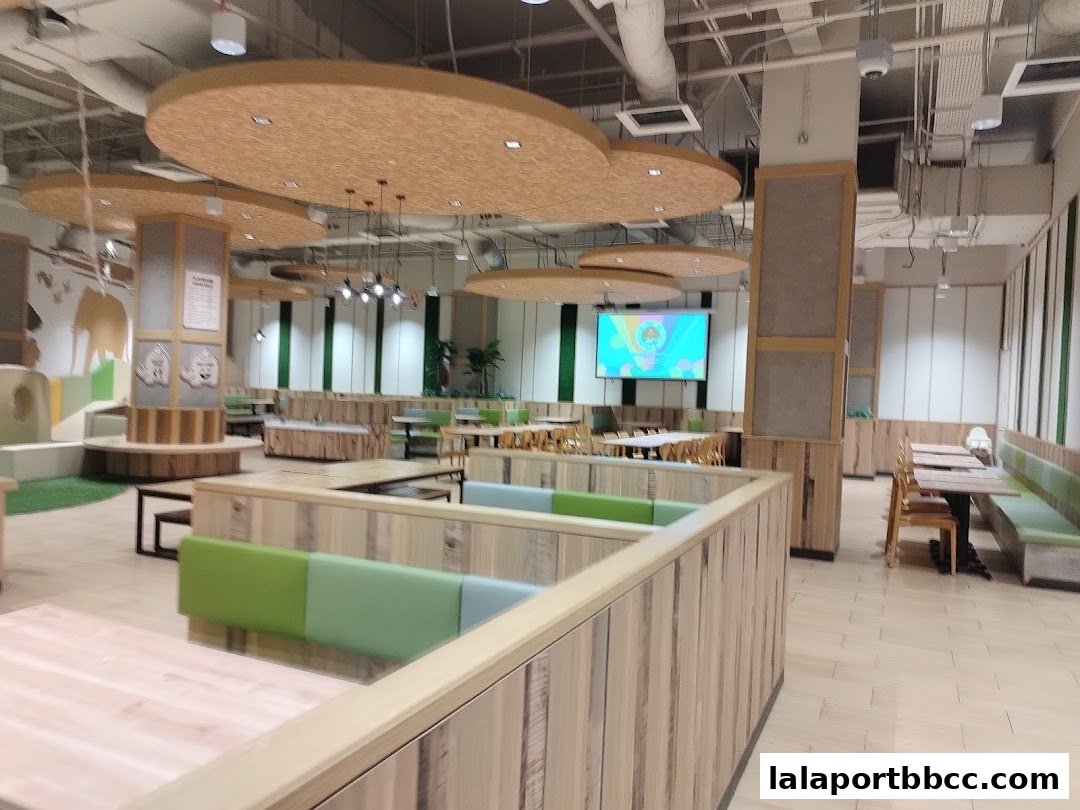 Spacious food court with modern wooden seating, green benches, and a large screen.