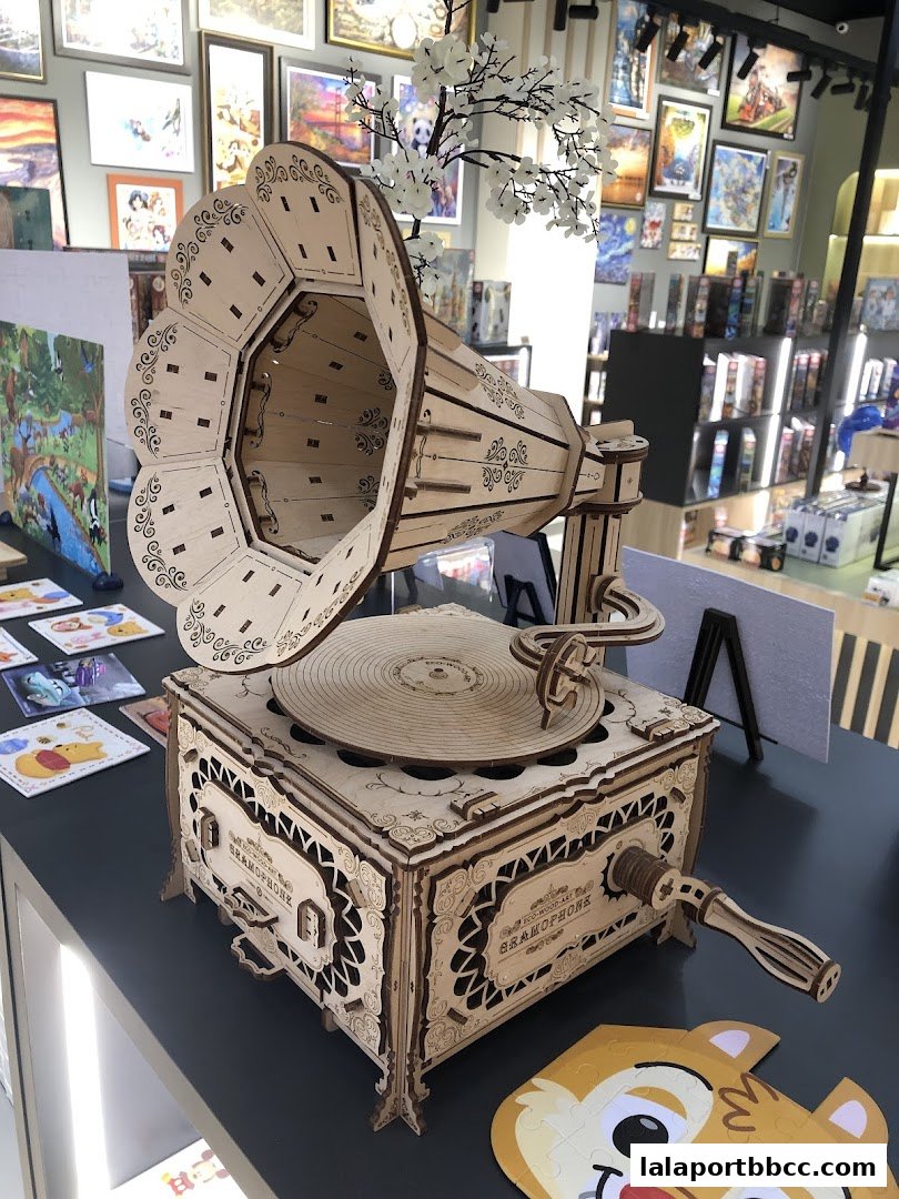 A detailed wooden model of a gramophone is displayed in a shop with puzzles and artwork in the background.