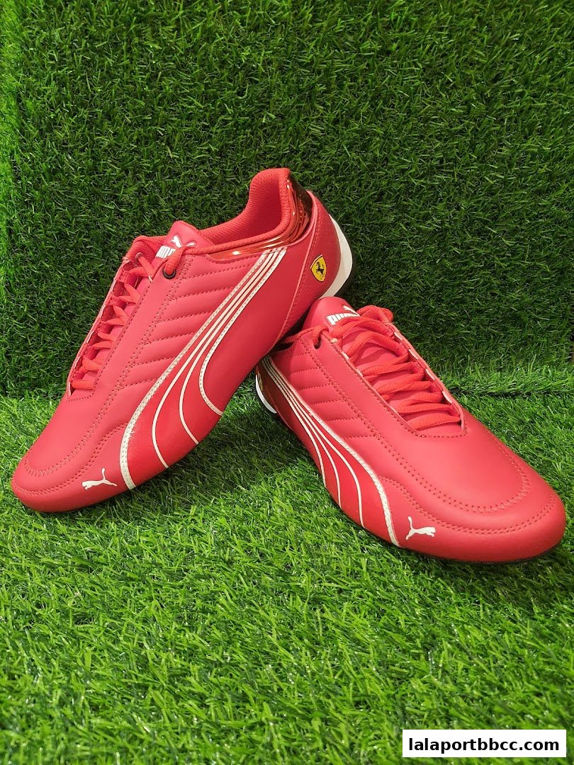 A pair of red Puma Ferrari racing shoes displayed on a green grassy surface.