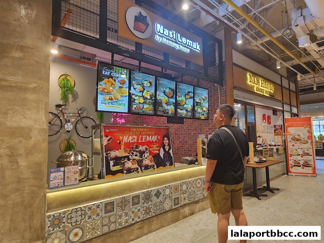 A man stands in front of a Nasi Lemak stall with digital menus displaying food items.