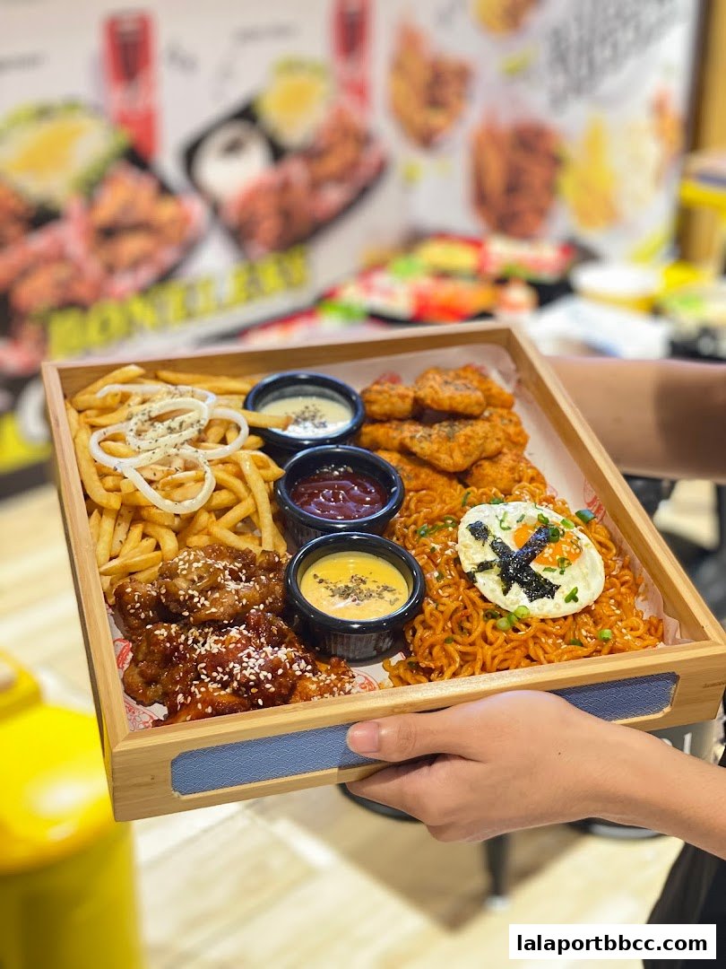A person holds a wooden tray filled with fried chicken, fries, dipping sauces, and spicy noodles.