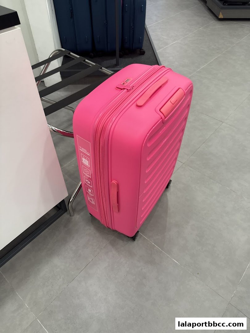 A bright pink suitcase is displayed in a retail store, with other luggage in the background.