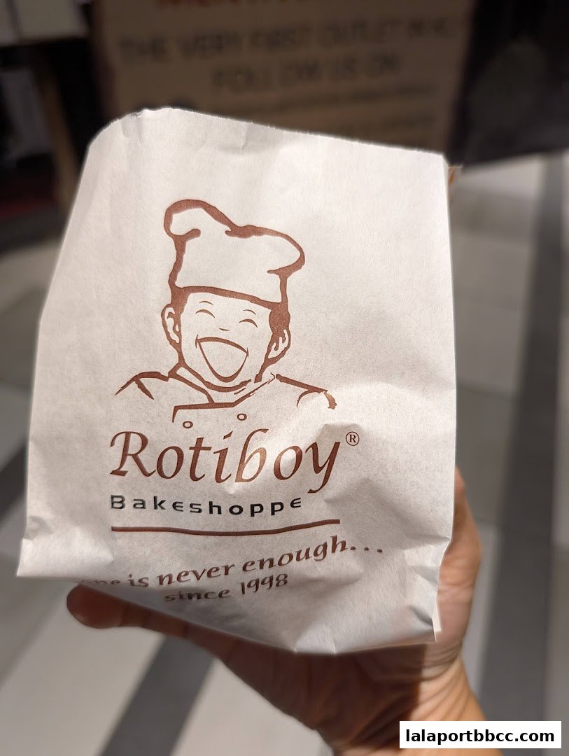 A hand holds a paper bag with the Rotiboy Bakeshoppe logo and slogan.