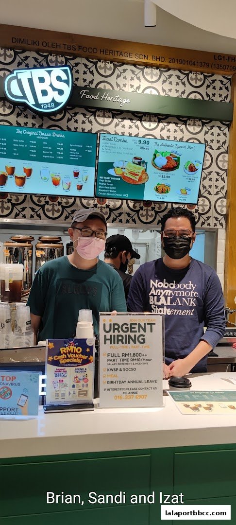 Two staff members stand behind a counter at TBS Food Heritage, with menus and a hiring sign visible.