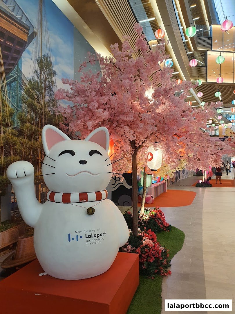 A large Maneki-neko statue and cherry blossom trees decorate a mall walkway.
