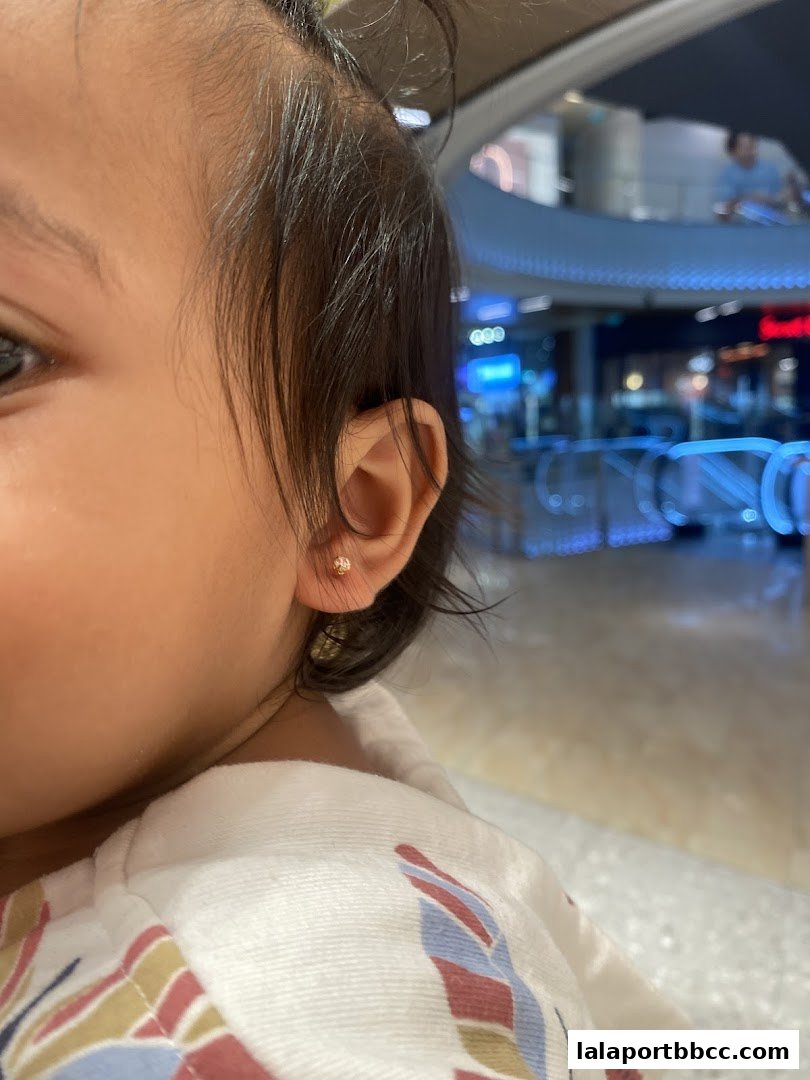 Close-up of a child's ear with a small stud earring, with a blurred mall background.