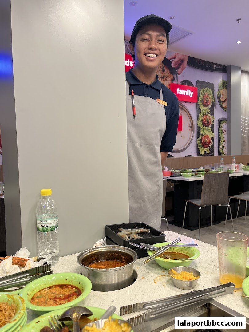 A smiling staff member stands behind a table with hot pot ingredients and condiments.