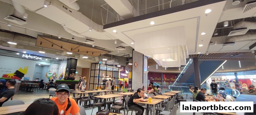 A busy cafeteria with people dining at tables and a view of escalators and other shops.