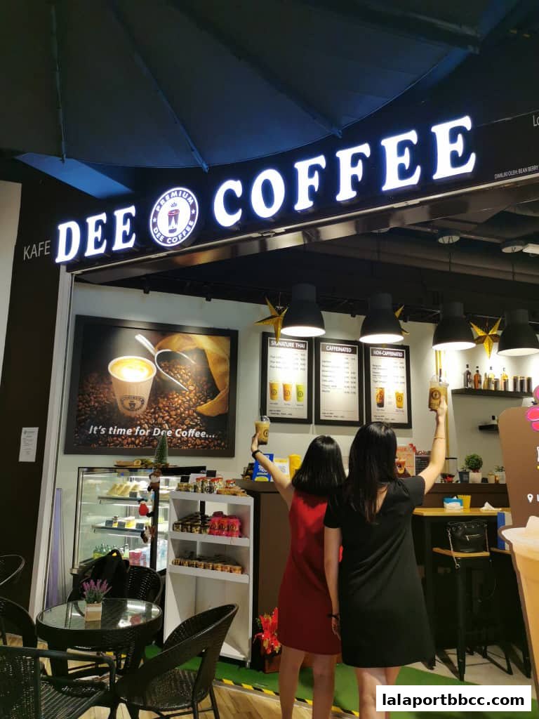 Two women raise coffee cups in front of a Dee Coffee shop with a large illuminated sign.