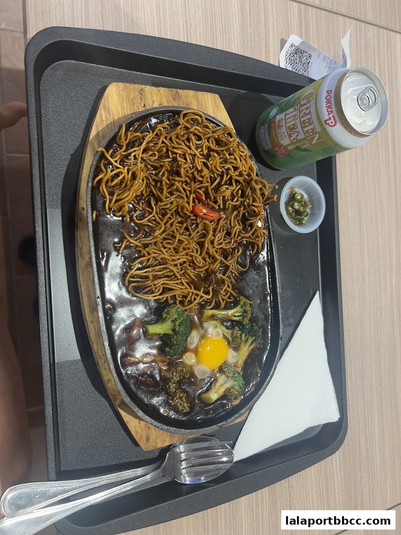 A sizzling plate of noodles with broccoli and egg, served with a can of green tea.