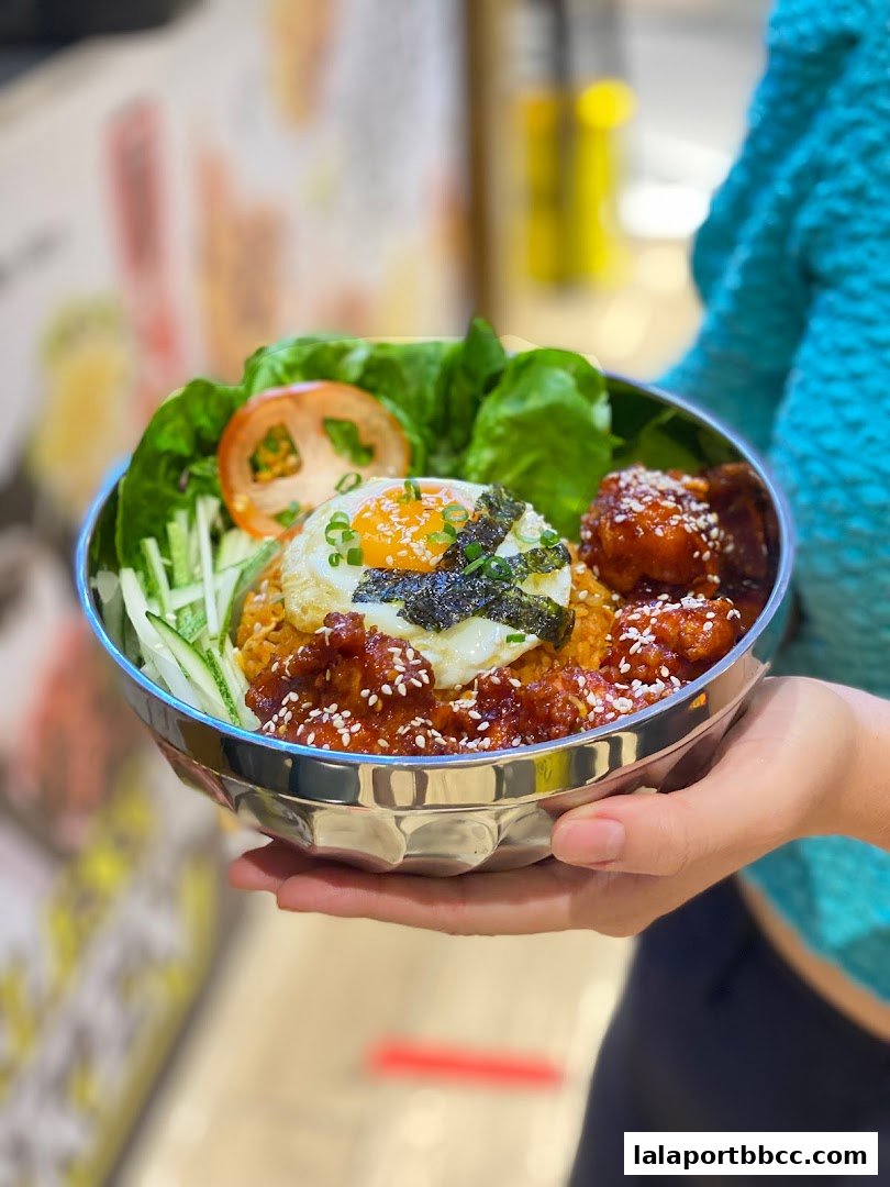 A person holds a bowl of Korean fried chicken with rice, a fried egg, and fresh vegetables.