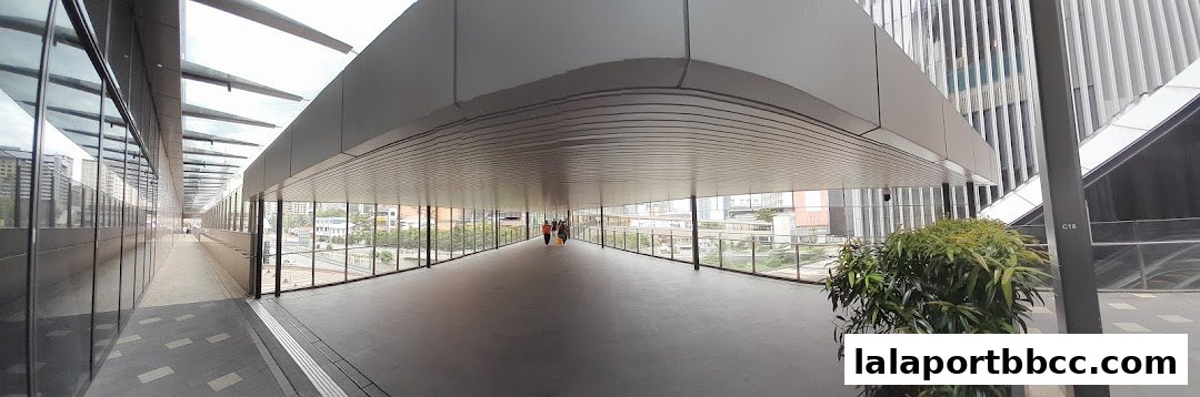 A panoramic view of a modern, covered pedestrian walkway connecting buildings, with glass walls and a view of the city.