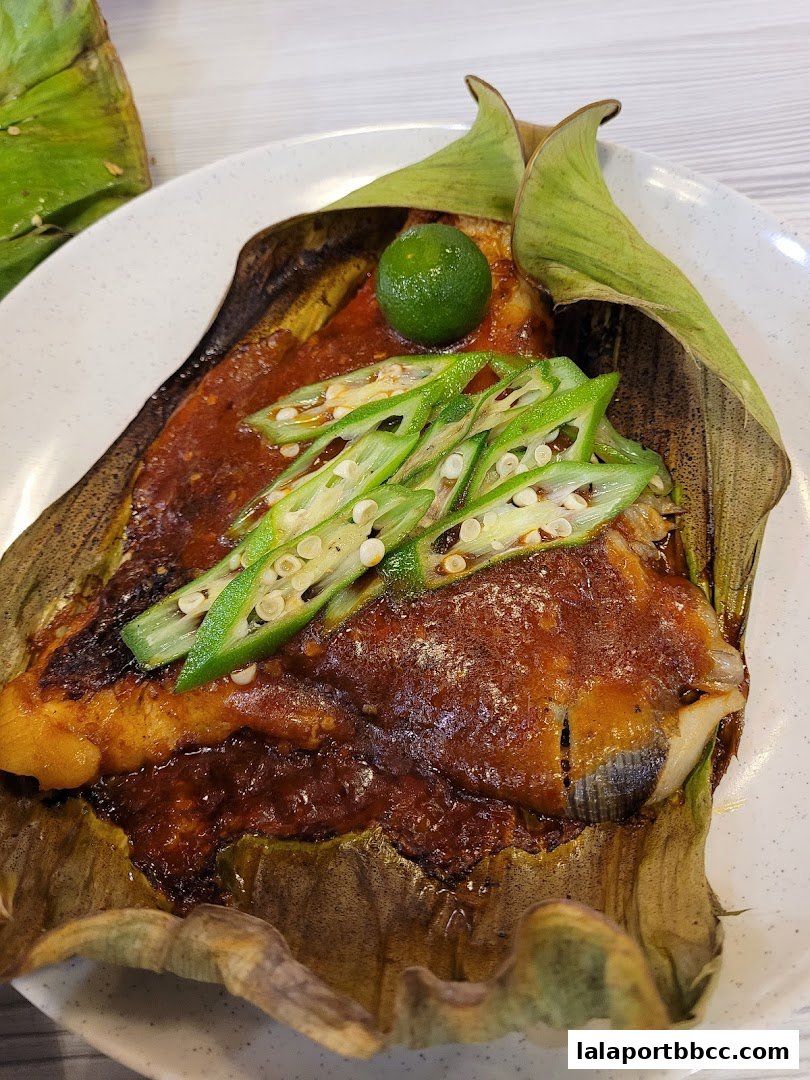 A close-up of grilled fish wrapped in banana leaves, topped with okra and a lime wedge.
