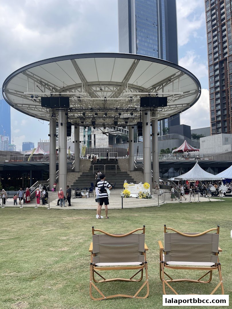An outdoor stage area with seating and people gathered, set against a backdrop of city buildings.