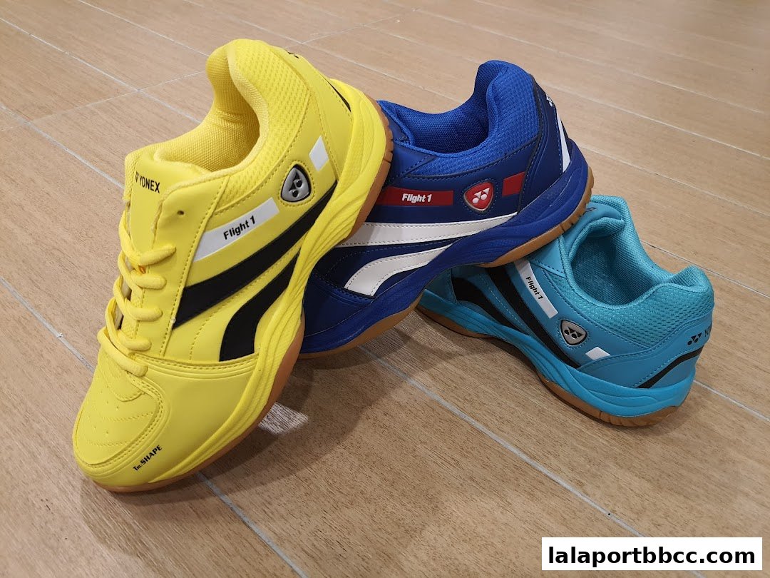 Three colorful Yonex badminton shoes, yellow, blue, and teal, are displayed on a wooden floor.