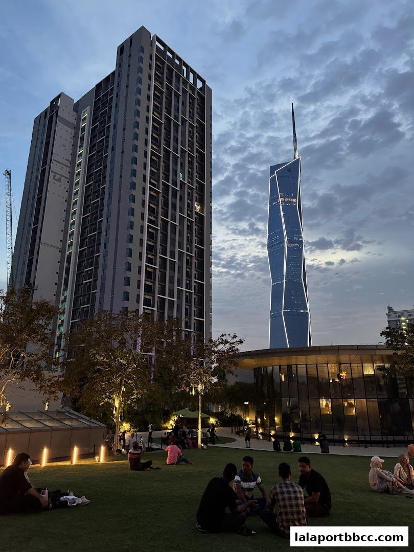 People relaxing on a lawn with modern buildings and a distinctive skyscraper in the background.
