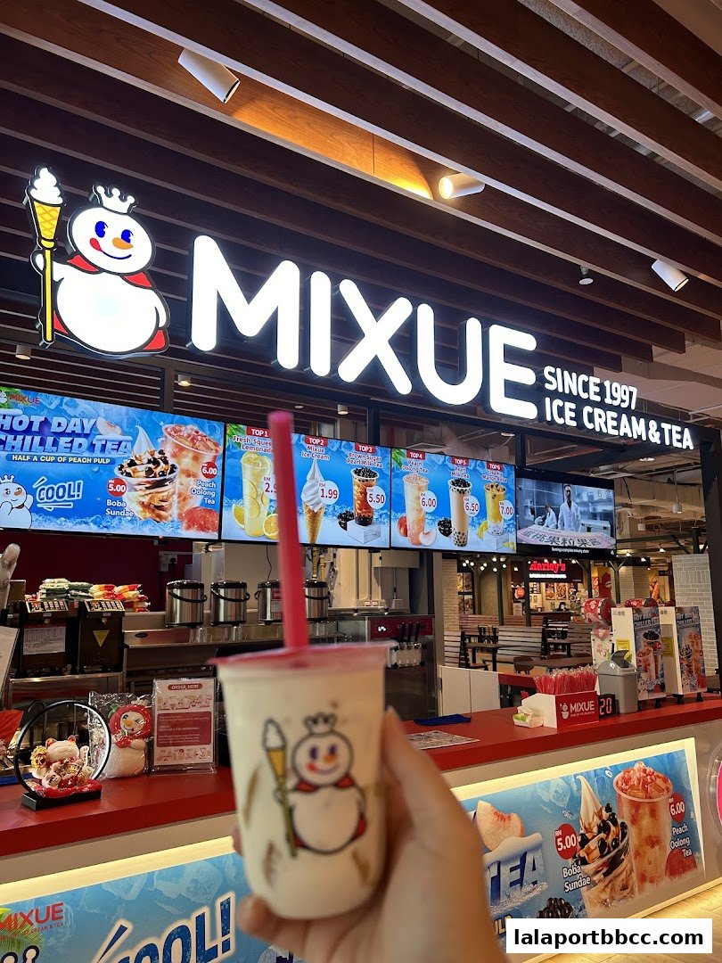 A hand holds a MIXUE drink in front of the shop's illuminated sign and menu.