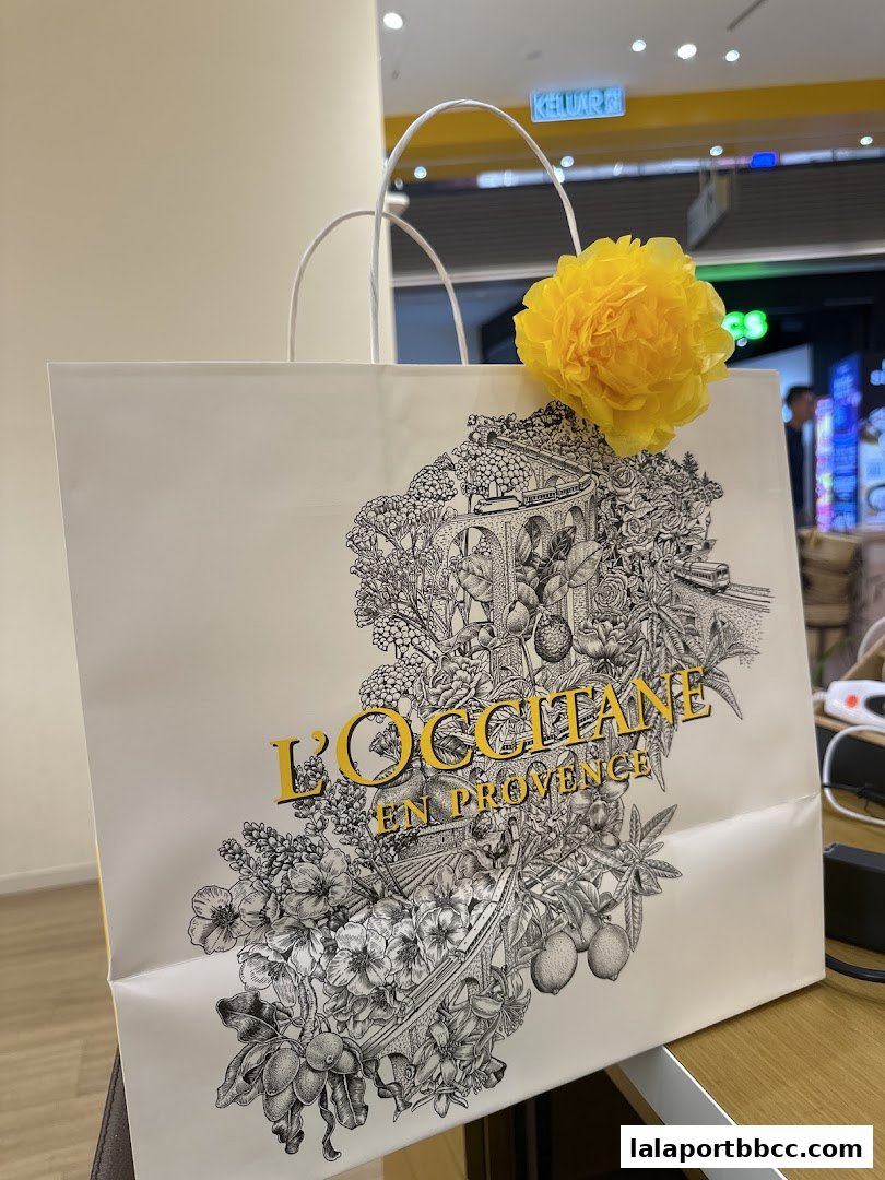 A shopping bag from L'Occitane En Provence decorated with intricate floral and architectural designs, topped with a yellow flower.