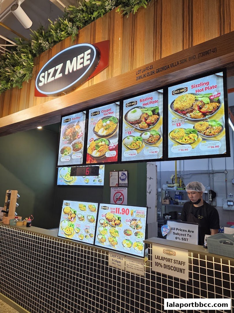 A food court counter with digital menus displaying various noodle dishes and a staff member.