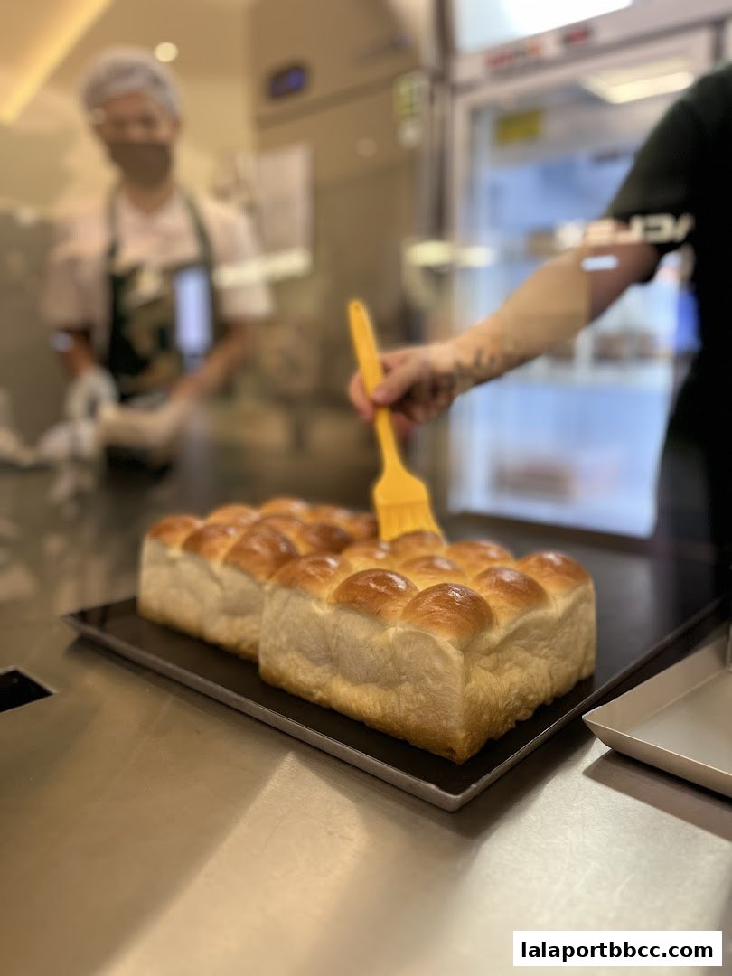 A baker is brushing glaze onto freshly baked bread rolls in a kitchen.