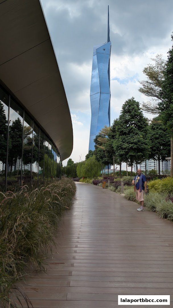 A person walks on a wooden pathway with lush greenery and a distinctive blue skyscraper in the background.