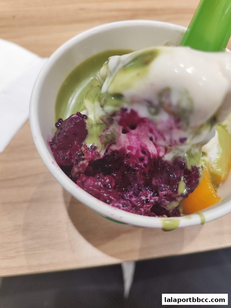 A cup of frozen yogurt with dragon fruit and other toppings, being scooped with a green spoon.