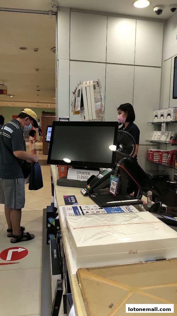 Customers at a checkout counter in an electronics store, with iPads and other products visible.