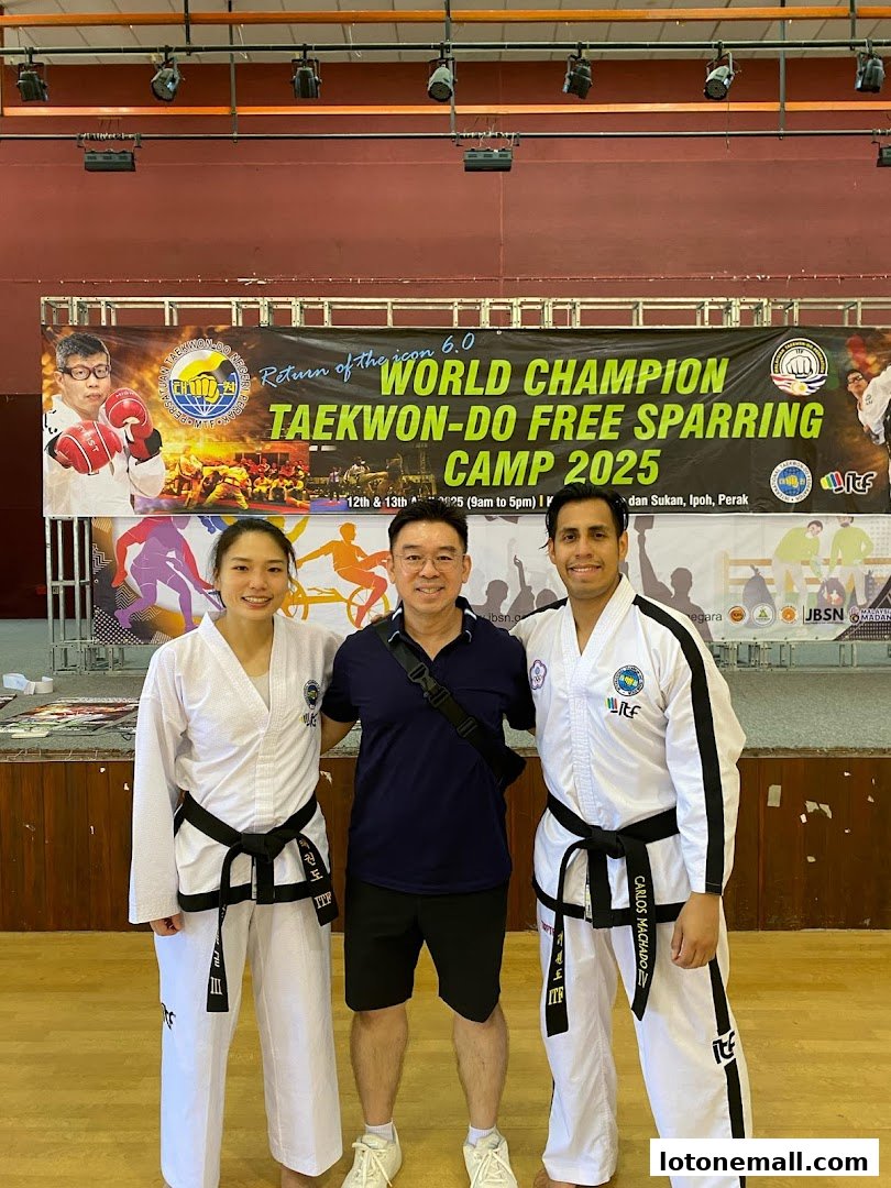 Three people in Taekwondo uniforms pose in front of a banner for a Taekwondo Free Sparring Camp.