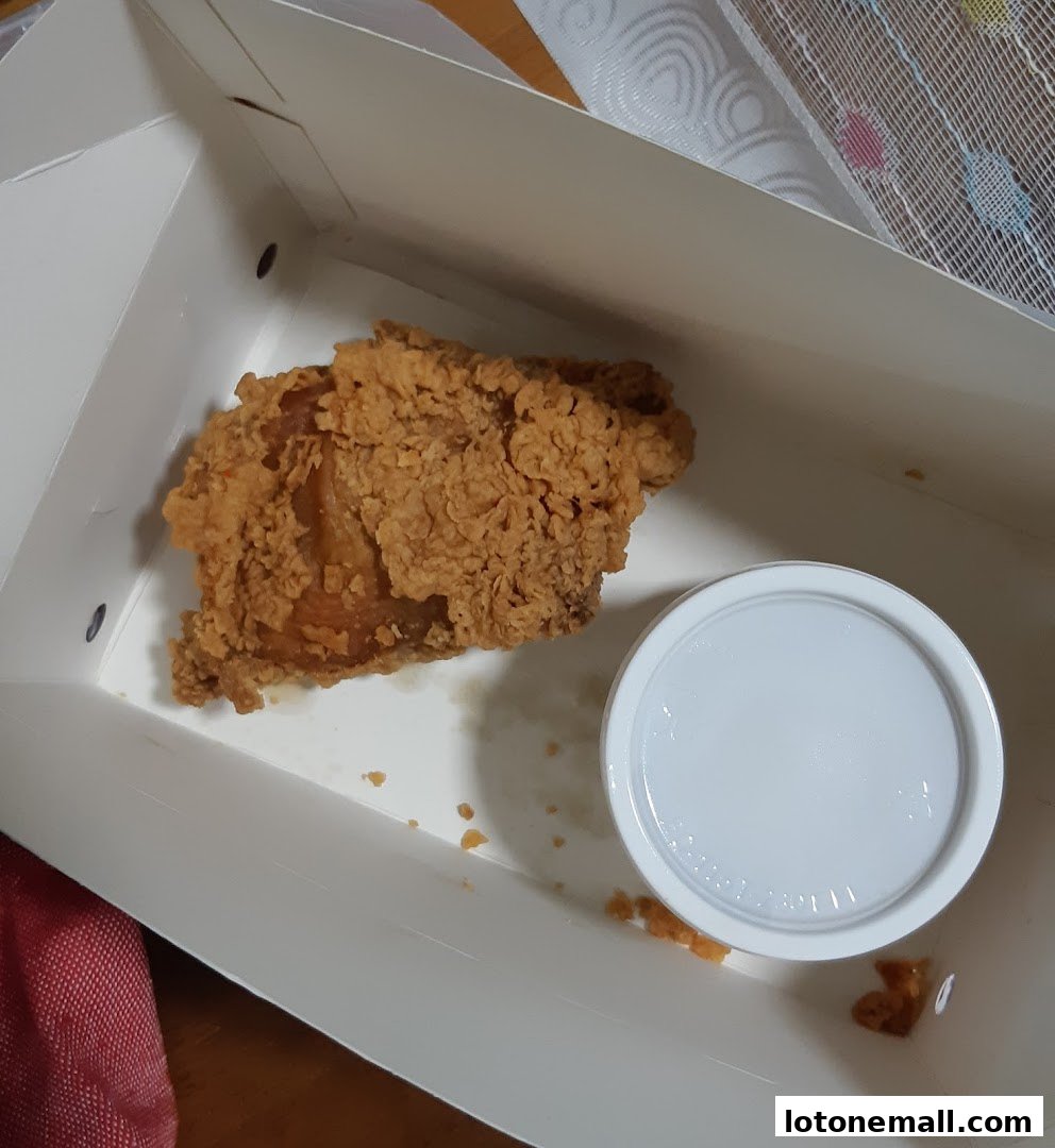 A piece of crispy fried chicken in a white takeaway box with a small container of sauce.