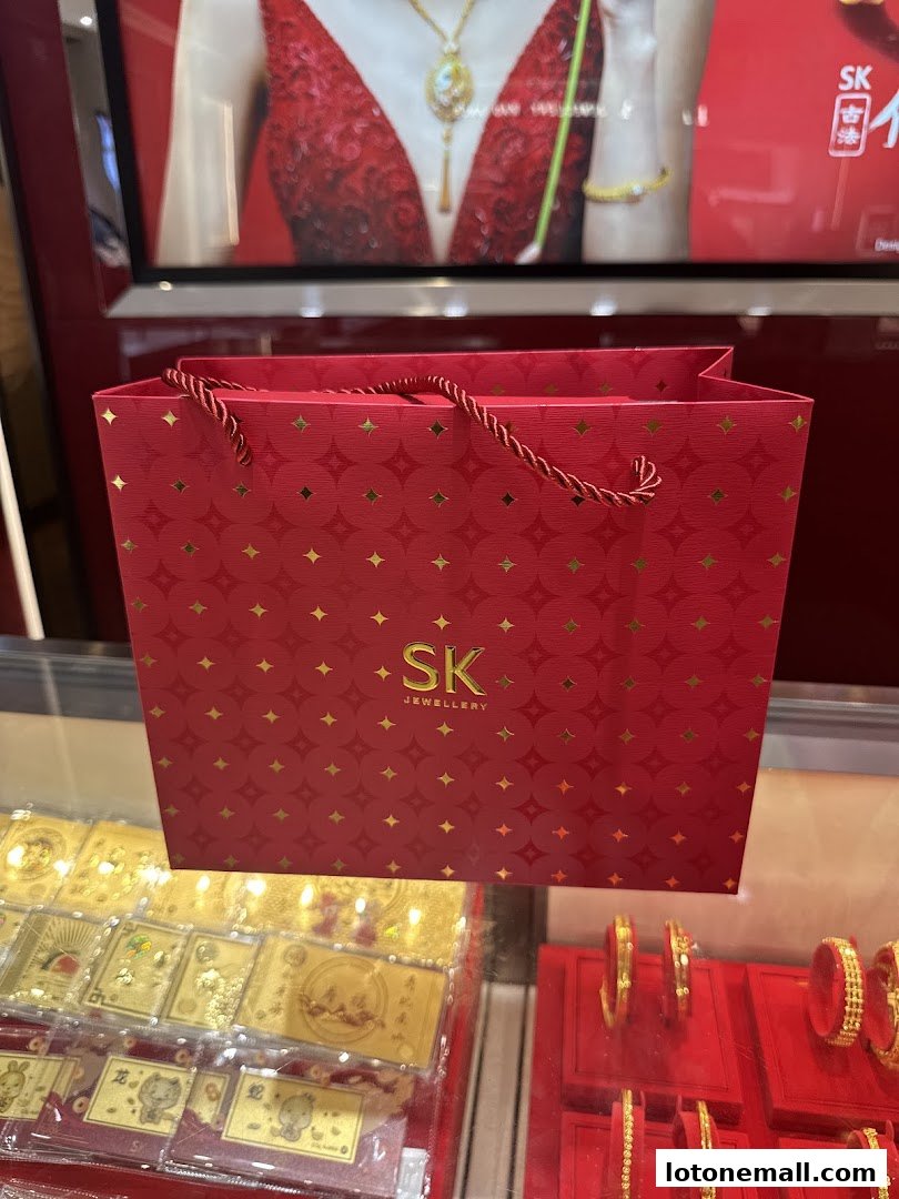 A red SK Jewellery shopping bag with gold patterns sits in front of display cases of gold jewelry.