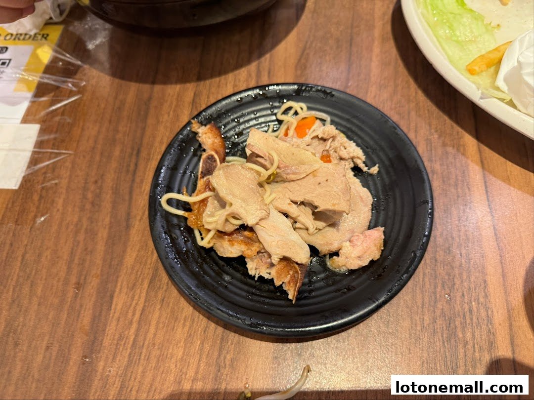 A black plate with sliced meat and noodles sits on a wooden table next to a salad.