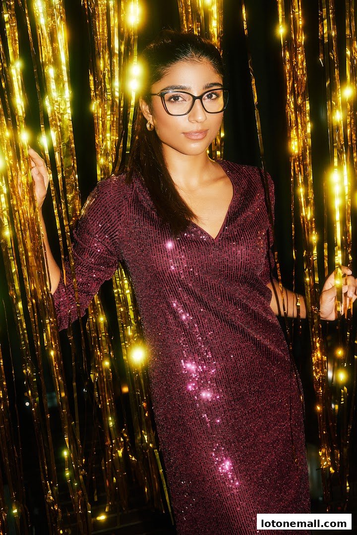 A woman wearing glasses and a sparkly dress poses against a backdrop of gold fringe.