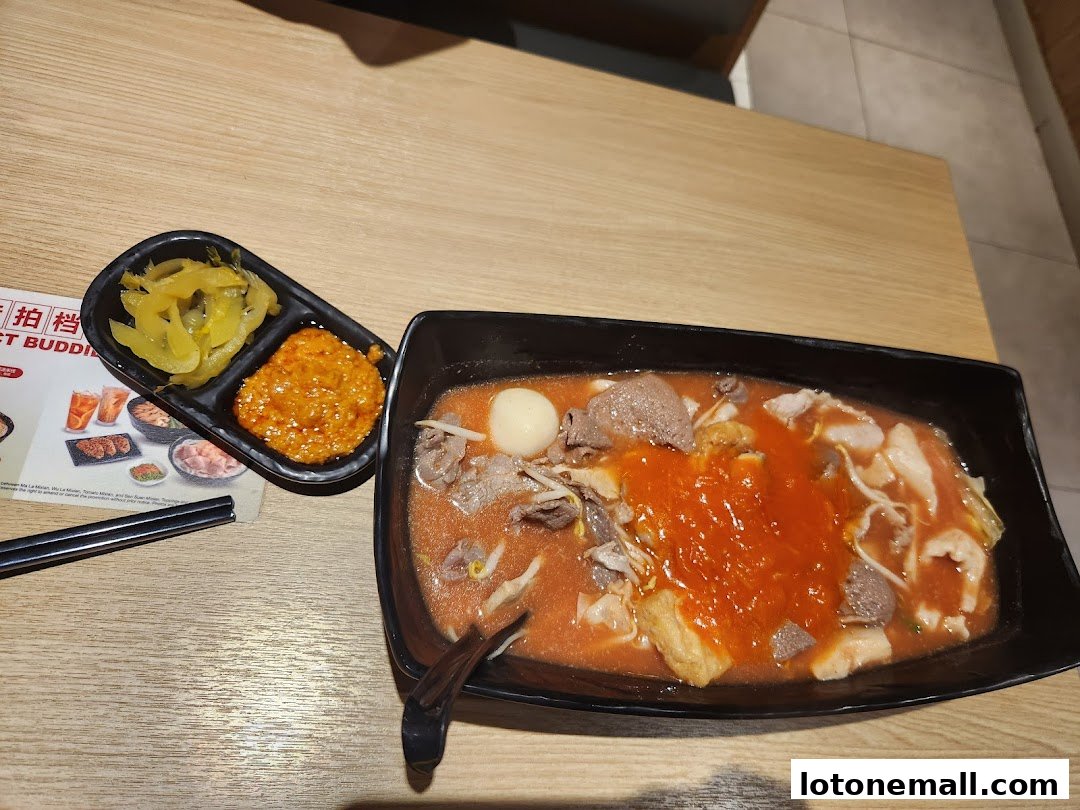 A bowl of spicy noodle soup with meat and pickled sides on a wooden table.
