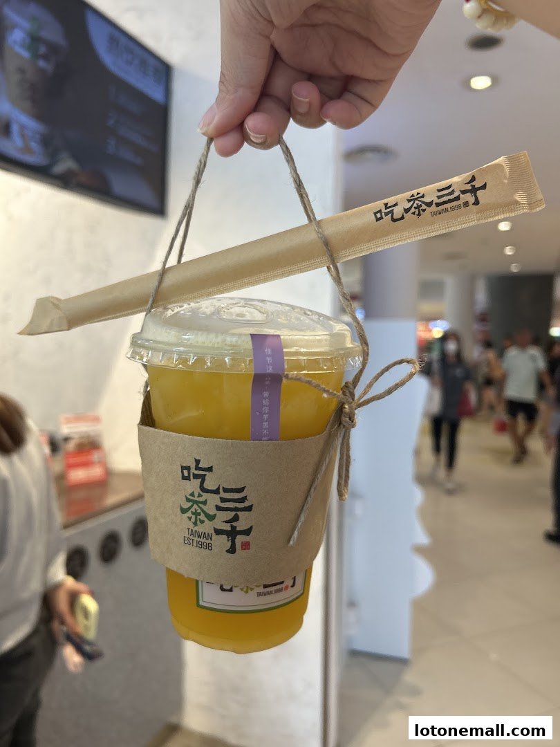 A hand holds a takeaway cup of orange beverage with a straw and branded sleeve.