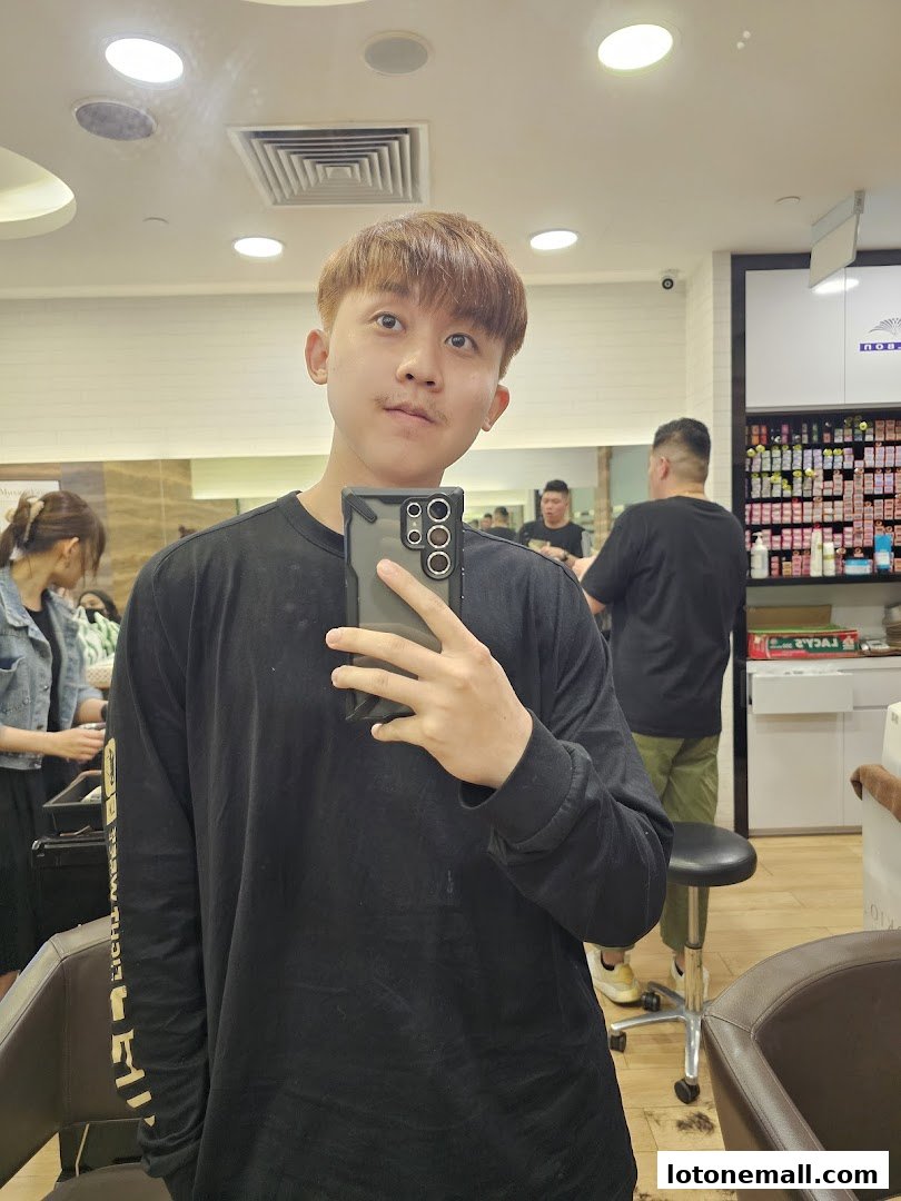 A man takes a selfie in a hair salon, with stylists and products visible in the background.