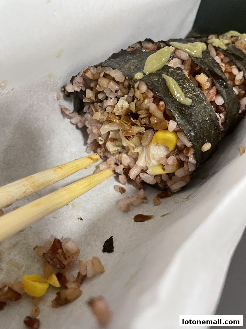 A close-up of a sushi roll with brown rice and corn, held by chopsticks.