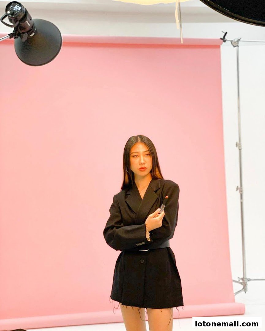 A woman in a black blazer holds a lipstick in front of a pink backdrop with studio lighting.