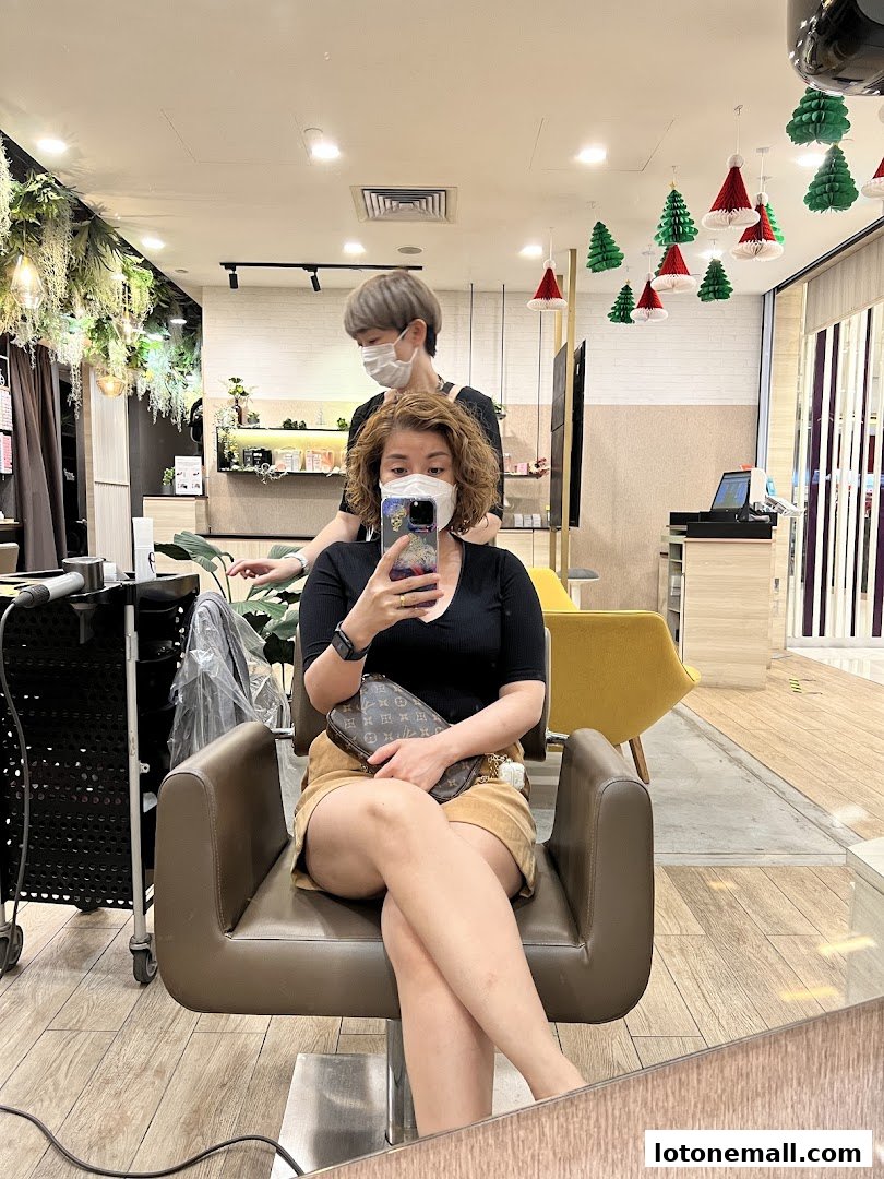 A woman sits in a salon chair while a stylist works on her hair, reflected in a mirror.