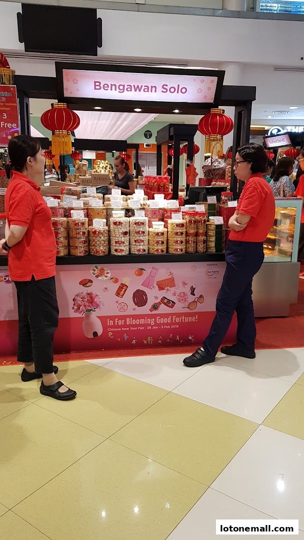 Two women in red shirts stand near a stall selling cookies and snacks, decorated for Chinese New Year.