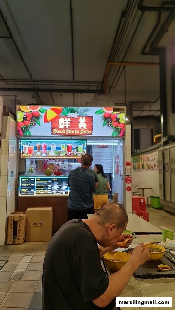 A vibrant juice stall named '鮮美 Fresh Fruits Juice' with customers and a person eating in the foreground.