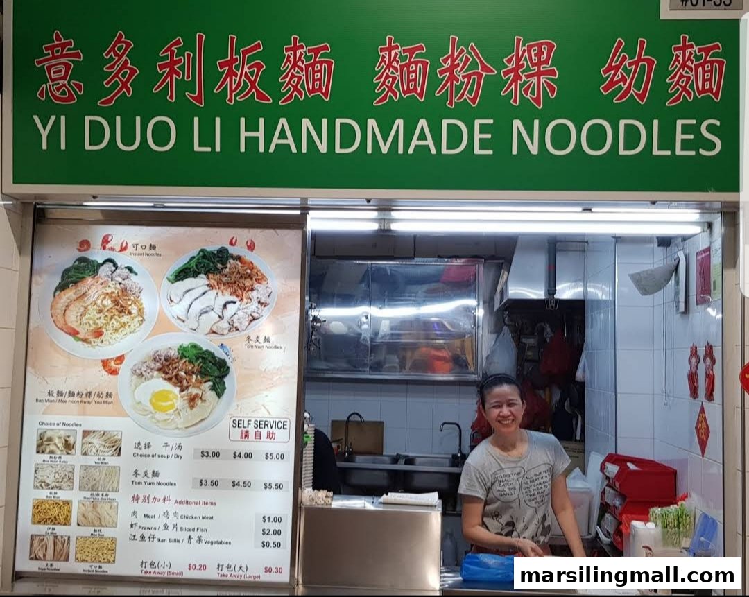 A hawker stall's menu board displaying various handmade noodle dishes and prices, with a staff member smiling inside.