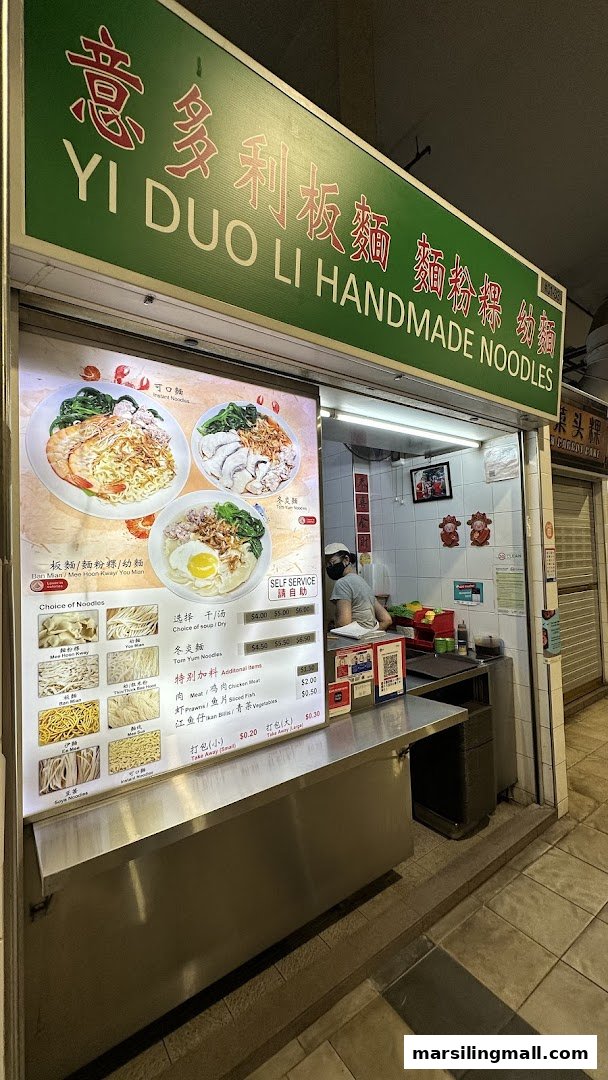 A brightly lit hawker stall front featuring a large menu board with various handmade noodle dishes and a chef inside.