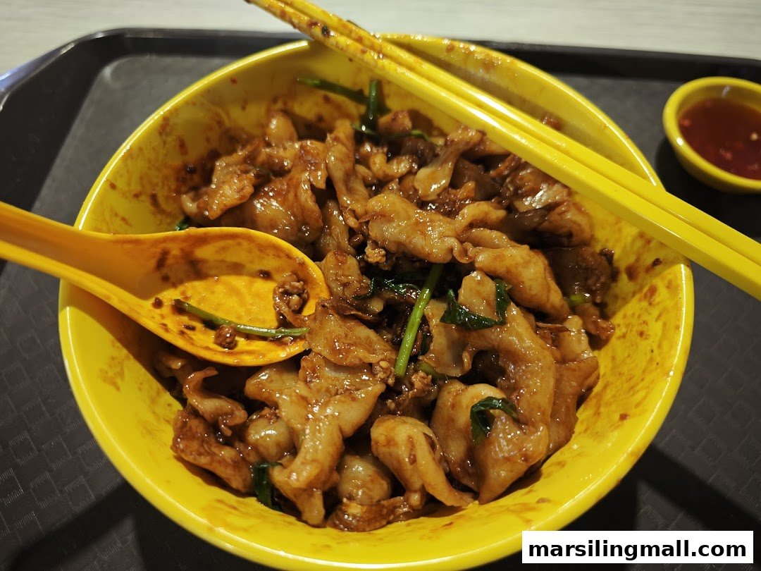 A vibrant yellow bowl filled with stir-fried noodles, meat, and greens, accompanied by a small dish of chili sauce.