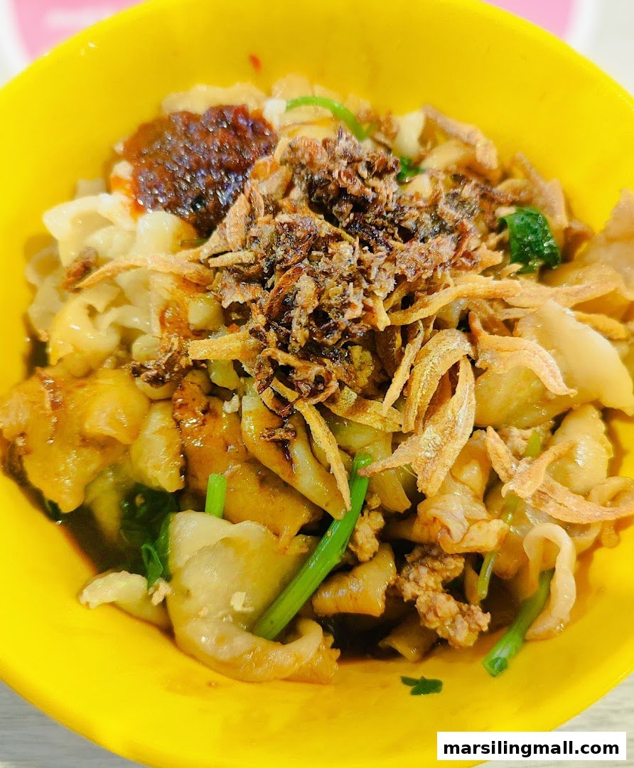 A vibrant yellow bowl filled with handmade noodles, topped with fried anchovies, crispy shallots, minced meat, and chili.