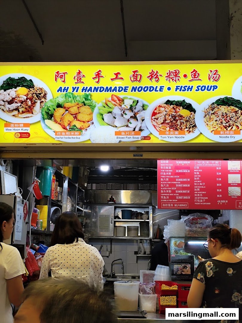 The image shows the bustling counter of Ah Yi Handmade Noodle Fish Soup with customers and a vibrant menu board.