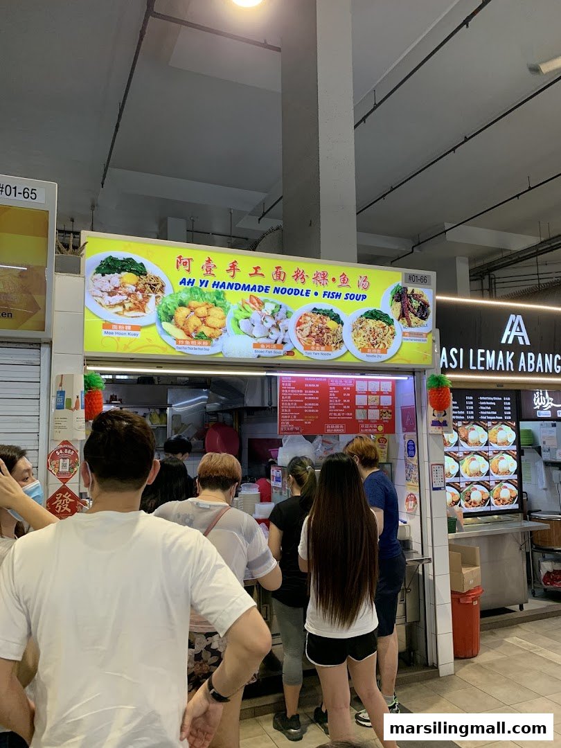 A bustling food stall, 'Ah Yi Handmade Noodle Fish Soup', with customers queuing for orders.